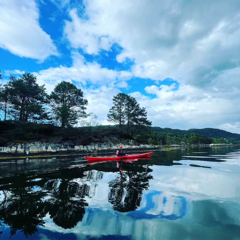 Rød kajakk på speilblank fjord med skog og skyer som speiler seg