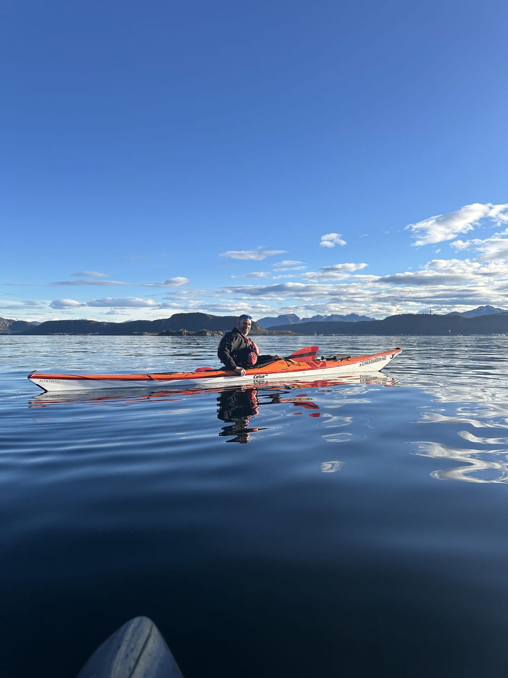 Padler i rød kajakk på blikkstille fjord under blå himmel